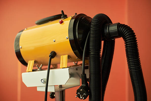 Industrial hot air blower standing on metal support, featuring yellow cylindrical body with attached black flexible hoses and visible power cord against plain orange background